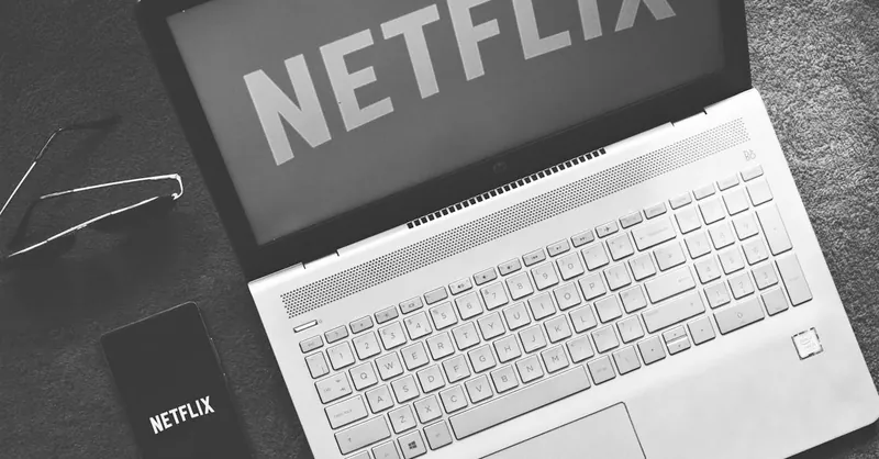 Monochrome image showcasing a laptop with Netflix on screen, smartphone, and sunglasses.